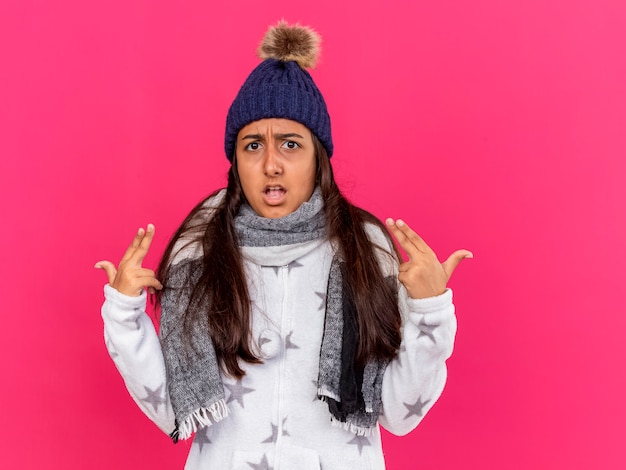 Unpleased young ill girl wearing winter hat with scarf points at herself isolated on pink background
