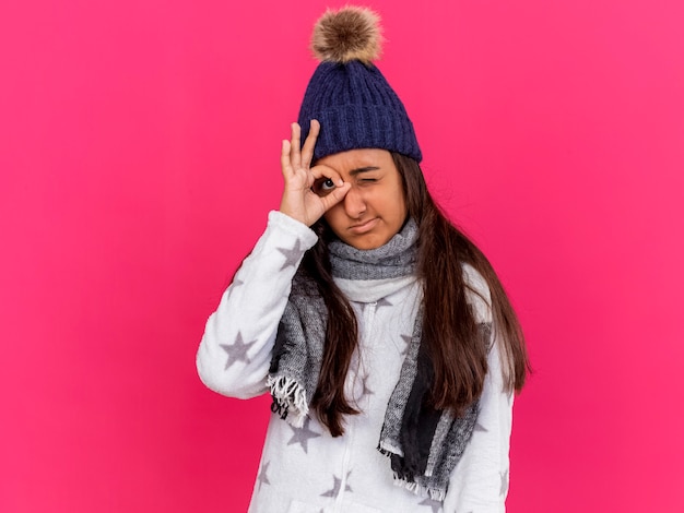 Недовольная молодая больная девушка в зимней шапке с шарфом показывает жест, изолированный на розовом