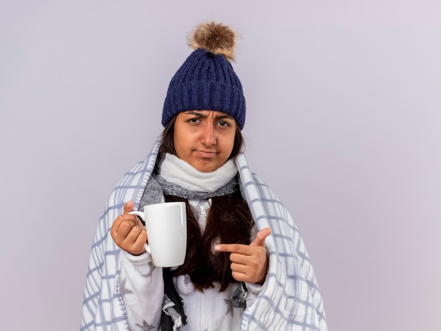 Unpleased young ill girl wearing winter hat with scarf wrapped in plaid holding and points at cup of tea