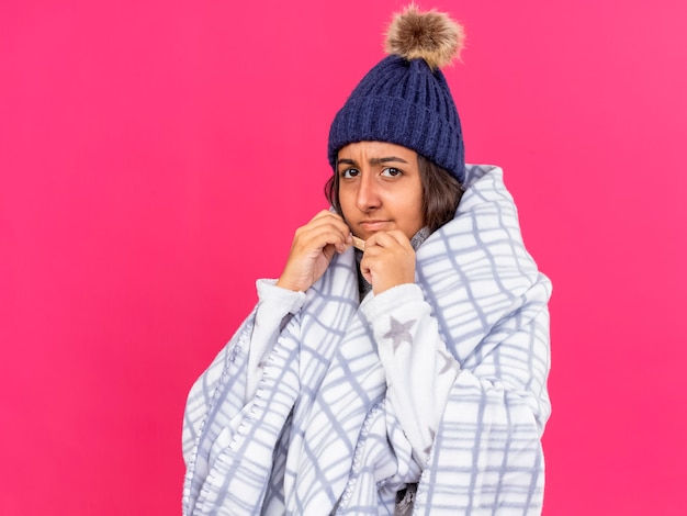 Недовольная молодая больная девушка в зимней шапке с шарфом, завернутым в плед, кладет пластырь на нос, изолированные на розовом фоне