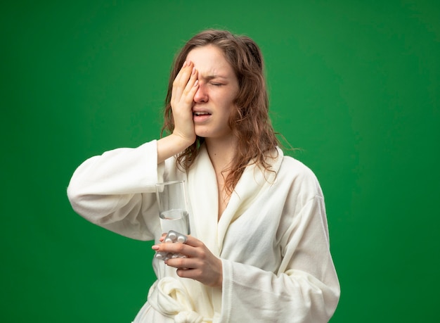 Foto gratuita giovane ragazza malata dispiaciuta con gli occhi chiusi che indossa una veste bianca che tiene il bicchiere d'acqua mettendo la mano sul viso isolato sul verde