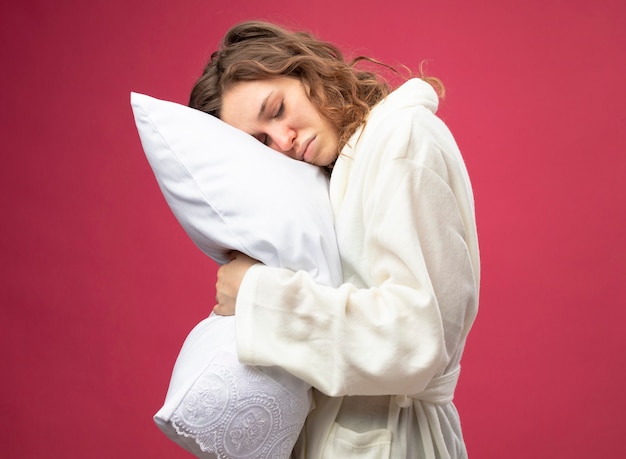 ピンクで隔離の白いローブ抱き枕を身に着けている目を閉じて不機嫌な若い病気の女の子