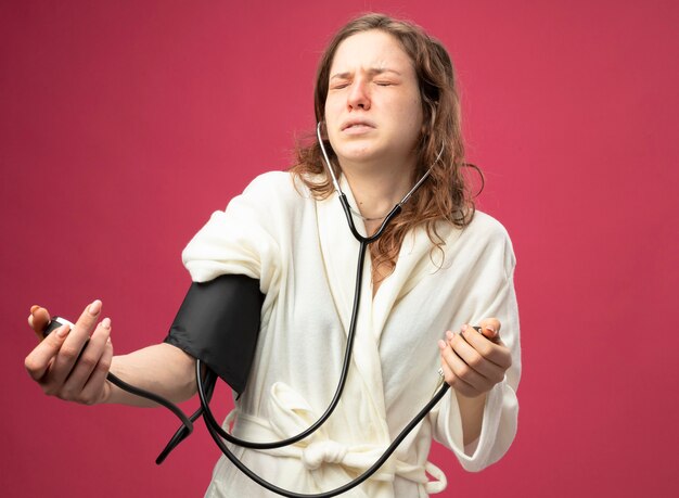 Unpleased young ill girl with closed eyes wearing white robe measuring her own pressure with sphygmomanometer isolated on pink