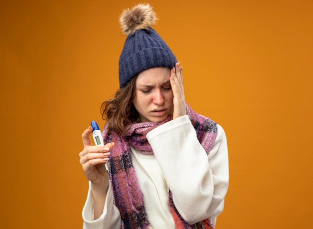 Unpleased young ill girl with closed eyes wearing white robe and winter hat with scarf holding thermometer putting hand on cheek isolated on orange