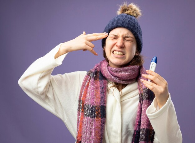 Unpleased young ill girl with closed eyes wearing white robe and winter hat with scarf holding thermometer showing suicide with pistol gesture isolated on purple