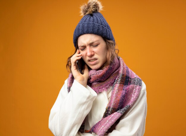 Unpleased young ill girl with closed eyes wearing white robe and winter hat with scarf speaks on phone isolated on orange wall