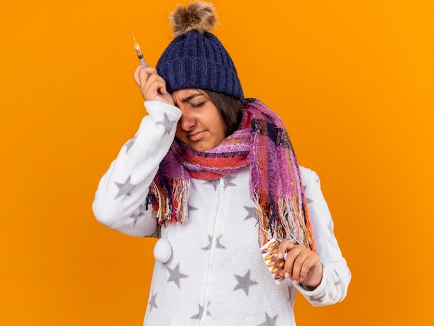 Unpleased young ill girl with closed eyes wearing winter hat with scarf holding pills with syringe and putting hand on forehead isolated on yellow background