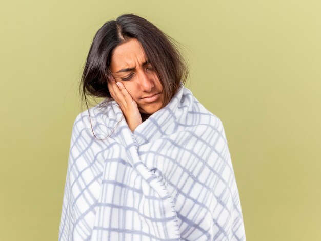 Unpleased young ill girl with closed eyes wrapped in plaid putting hand on cheek isolated on olive green background