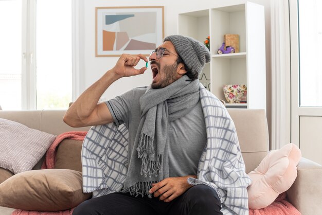 unpleased young ill man in optical glasses wrapped in plaid with scarf around his neck wearing winter hat taking medical pill sitting on couch at living room