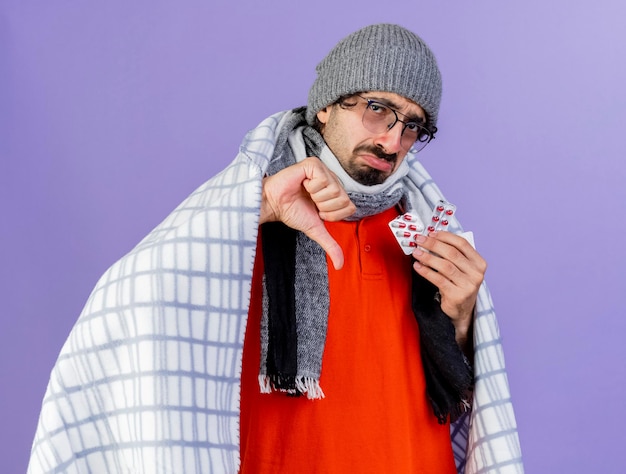 Foto gratuita giovane uomo malato dispiaciuto che indossa occhiali cappello invernale e sciarpa avvolti in un plaid che tiene confezioni di capsule guardando davanti che mostra il pollice verso il basso isolato sul muro viola