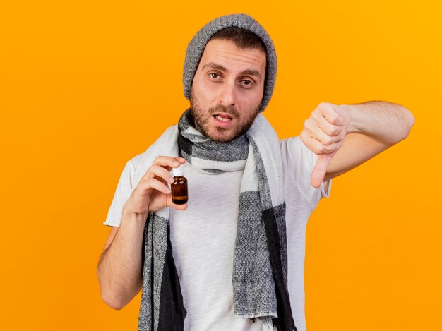 冬の帽子とスカーフを身に着けている不機嫌な若い病気の男が黄色の背景で隔離の親指を示すガラス瓶に薬を保持