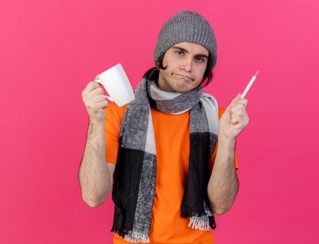 Unpleased young ill man wearing winter hat with scarf holding cup of tea with thermometer isolated on pink