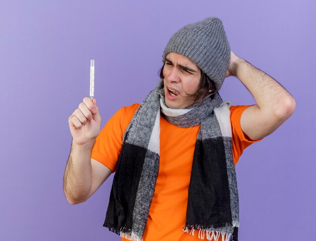 紫色の背景で隔離の頭に手を置いてスカーフを保持し、温度計を見て冬の帽子をかぶって不機嫌な若い病気の男