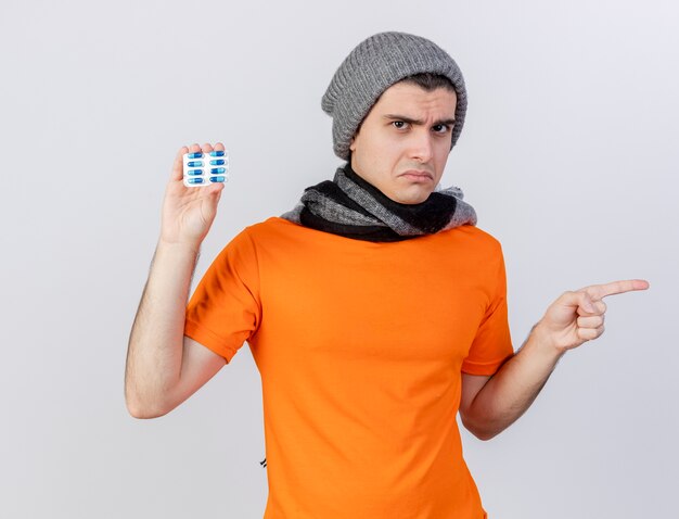 Unpleased young ill man wearing winter hat with scarf holding pills and points at side isolated on white