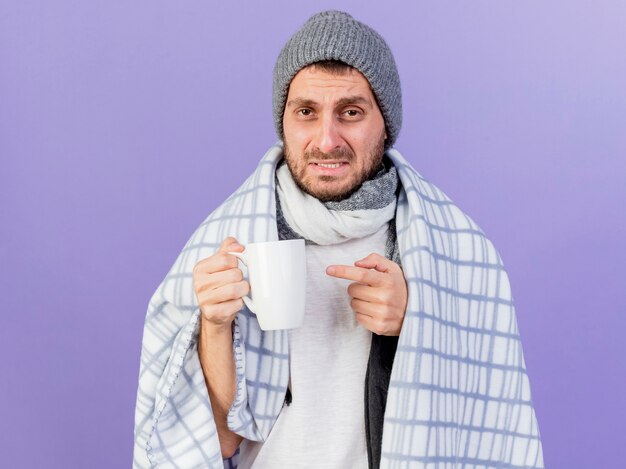 Unpleased young ill man wearing winter hat with scarf holding and points at cup of tea isolated on purple background