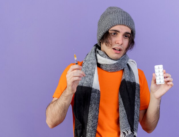 unpleased young ill man wearing winter hat with scarf holding syringe with pills isolated on purple