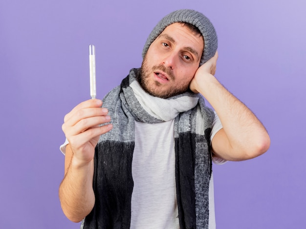 Unpleased young ill man wearing winter hat with scarf holding thermometer and putting hand on head isolated on purple background