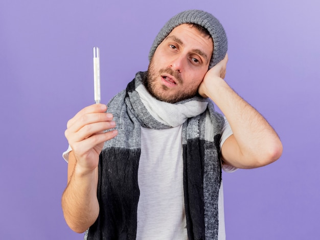 温度計を保持し、紫色の背景で隔離の頭に手を置くスカーフと冬の帽子をかぶって不機嫌な若い病気の男
