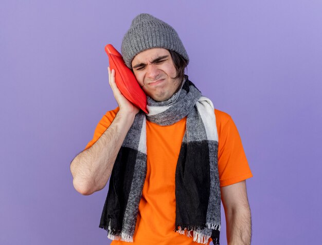unpleased young ill man wearing winter hat with scarf putting hot water bag on cheek isolated on purple