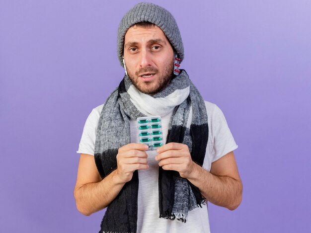 Недовольный молодой больной человек в зимней шапке с шарфом кладет таблетки под шляпу и держит таблетки, изолированные на фиолетовом фоне