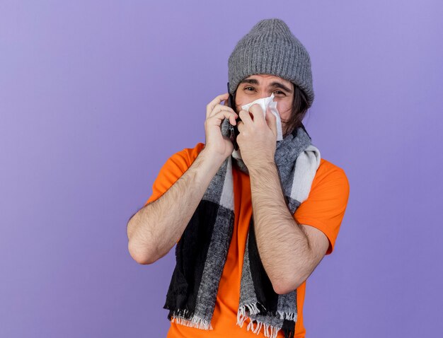 Недовольный молодой больной в зимней шапке с шарфом разговаривает по телефону и вытирает нос салфеткой, изолированной на фиолетовом фоне