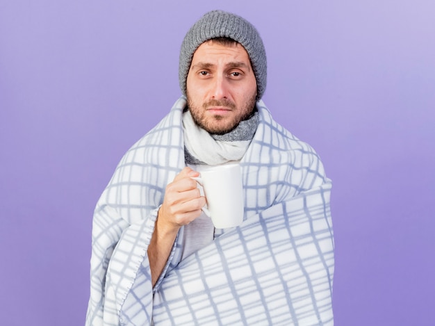 Unpleased young ill man wearing winter hat with scarf wrapped in plaid and holding cup uf tea isolated on purple background