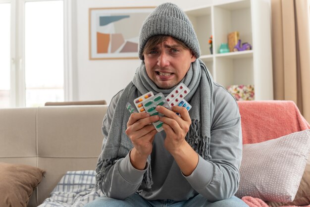 unpleased young ill man with scarf around neck wearing winter hat holding medicine blister packs sitting on couch at living room