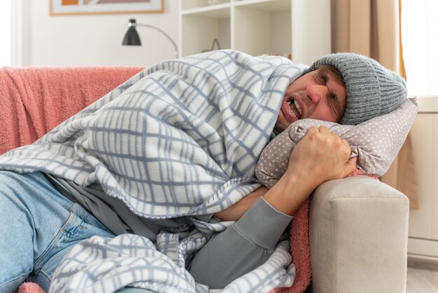 Unpleased young ill slavic man with scarf around neck wearing winter hat wrapped in plaid lying on couch at living room