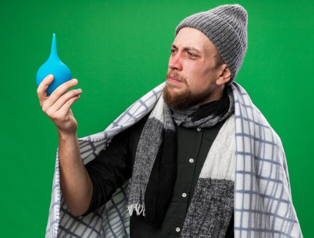 Unpleased young ill slavic man with scarf around neck wrapped in plaid wearing winter hat holding and looking at enema isolated on green wall with copy space