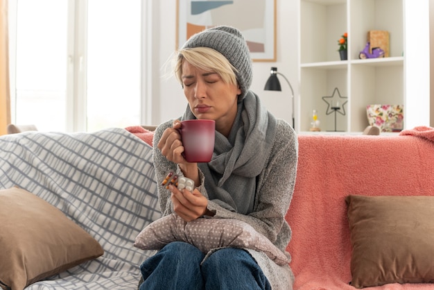 Unpleased young ill slavic woman with scarf around her neck wearing winter hat holding cup and medicine blister packs sitting on couch at living room