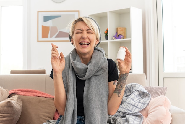 Unpleased young ill slavic woman with scarf around her neck wearing winter hat holding medicine bottle and crossing fingers sitting on couch with closed eyes at living room