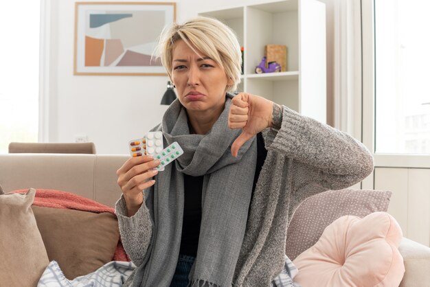 unpleased young ill slavic woman with scarf around her neck wrapped in plaid holding medicine blister packs and thumbing down sitting on couch at living room