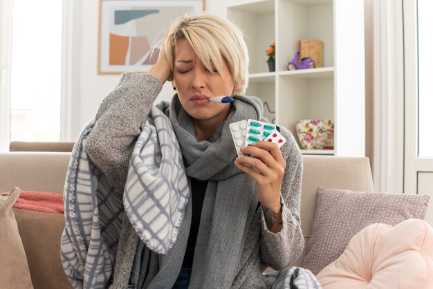 unpleased young ill slavic woman with scarf around her neck wrapped in plaid measuring her temperature with thermometer and holding medicine blister packs sitting on couch at living room