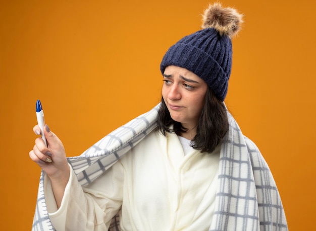 Foto gratuita giovane donna malata dispiaciuta che indossa il cappello invernale della veste avvolto in plaid che tiene e guardando la parte anteriore isolata sulla parete arancione