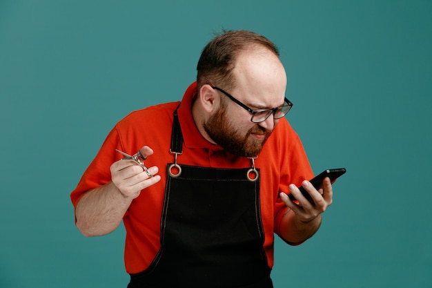 Unpleased young male barber wearing glasses red shirt and barber apron holding scissors and mobile phone looking at phone isolated on blue background