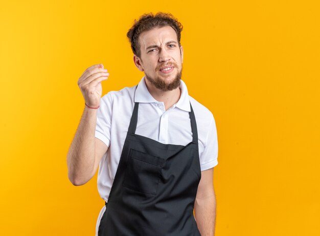 Unpleased young male barber wearing uniform showing tip gesture isolated on yellow background