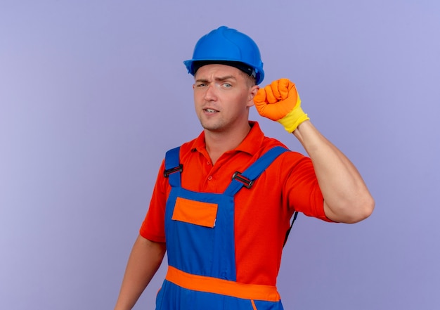 Unpleased young male builder wearing uniform and safety helmet in gloves raising fist on purple