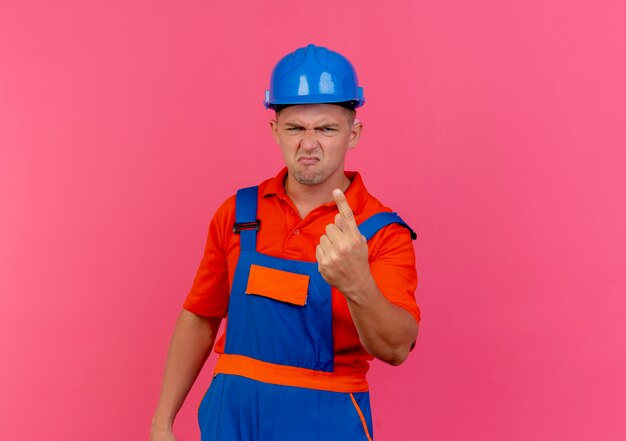 Unpleased young male builder wearing uniform and safety helmet showing one on pink