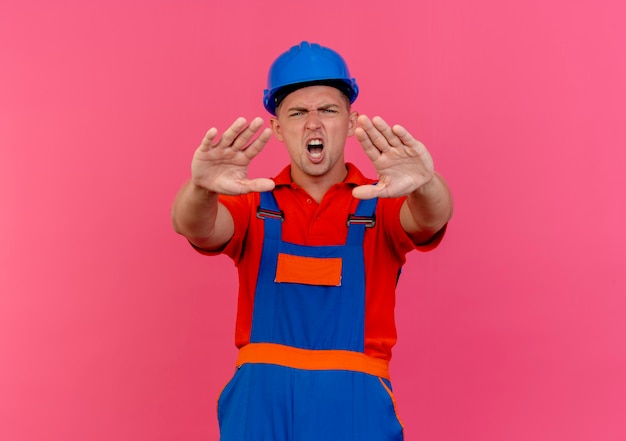 Unpleased young male builder wearing uniform and safety helmet showing stop gesture on pink