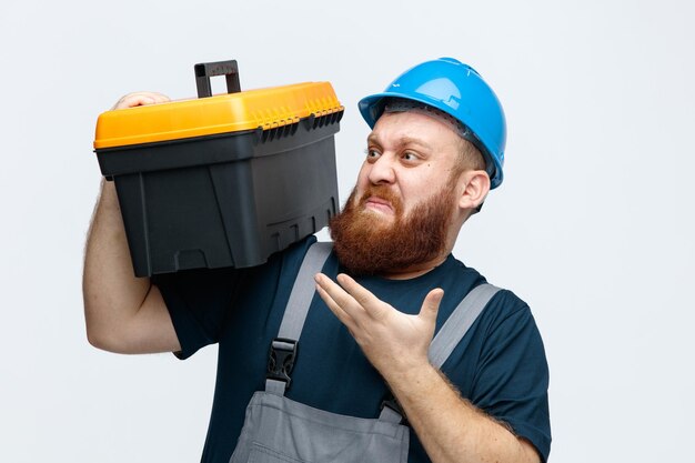 Unpleased young male construction worker wearing safety helmet and uniform holding tool box on shoulder looking at it showing empty hand isolated on white background