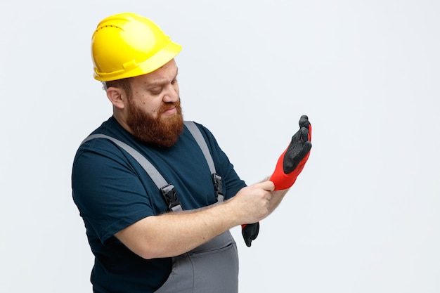 Unpleased young male construction worker wearing safety helmet and uniform standing in profile view wearing safety gloves looking at them isolated on white background