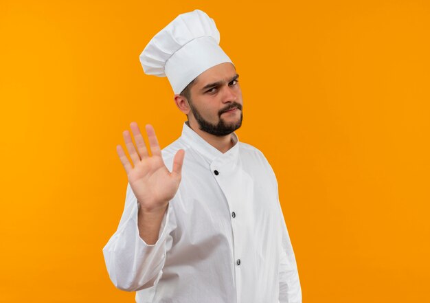 Unpleased young male cook in chef uniform gesturing stop isolated on orange space