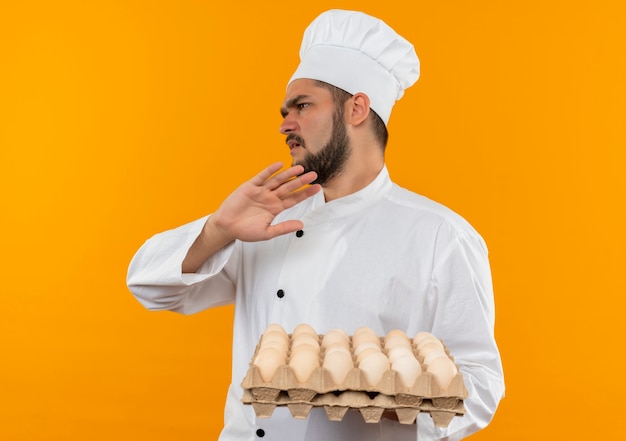 Unpleased young male cook in chef uniform holding carton of eggs looking at side and gesturing no isolated on orange space