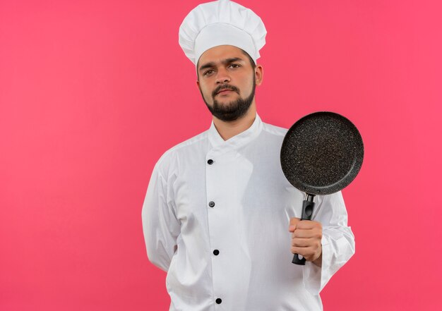 Unpleased young male cook in chef uniform holding frying pan with one hand behind back isolated on pink space 