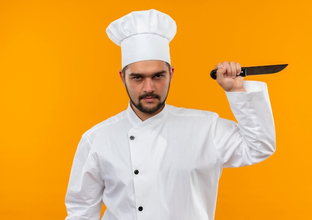 Unpleased young male cook in chef uniform holding knife isolated on orange space