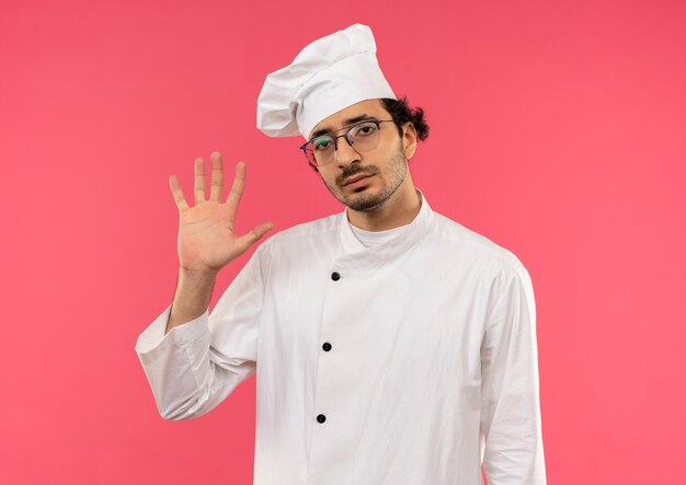 Unpleased young male cook wearing chef uniform and glasses showing stop gesture isolated on pink wall