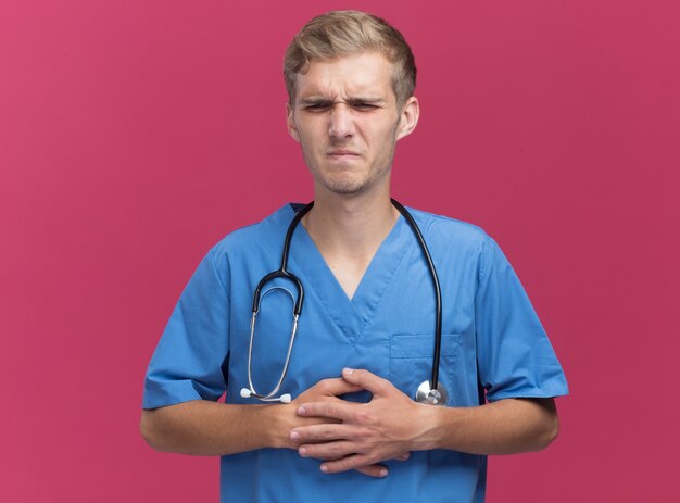 Unpleased young male doctor wearing doctor uniform with stethoscope putting hands on aching stomach isolated on pink wall