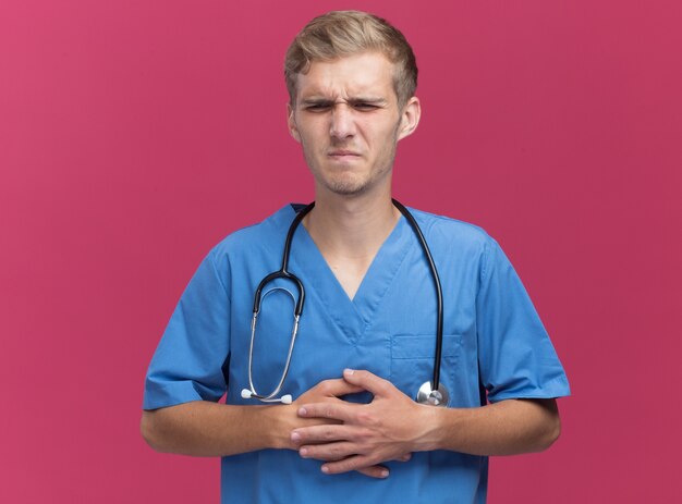ピンクの壁に隔離された痛む胃に手を置く聴診器で医者の制服を着て不機嫌な若い男性医師