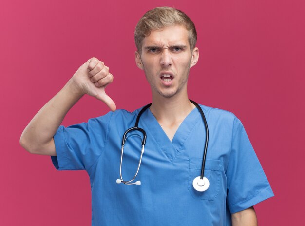 Недовольный молодой мужчина-врач в униформе врача со стетоскопом, показывающий большой палец вниз на розовой стене