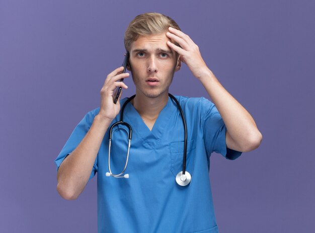 Unpleased young male doctor wearing doctor uniform with stethoscope speaks on phone putting hand on forehead isolated on blue wall